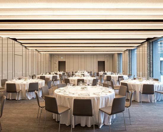 Configuration banquet dans une salle de réception avec couverts, nappes blanches et chaises à des tables rondes, portes d'entrée et grandes fenêtres